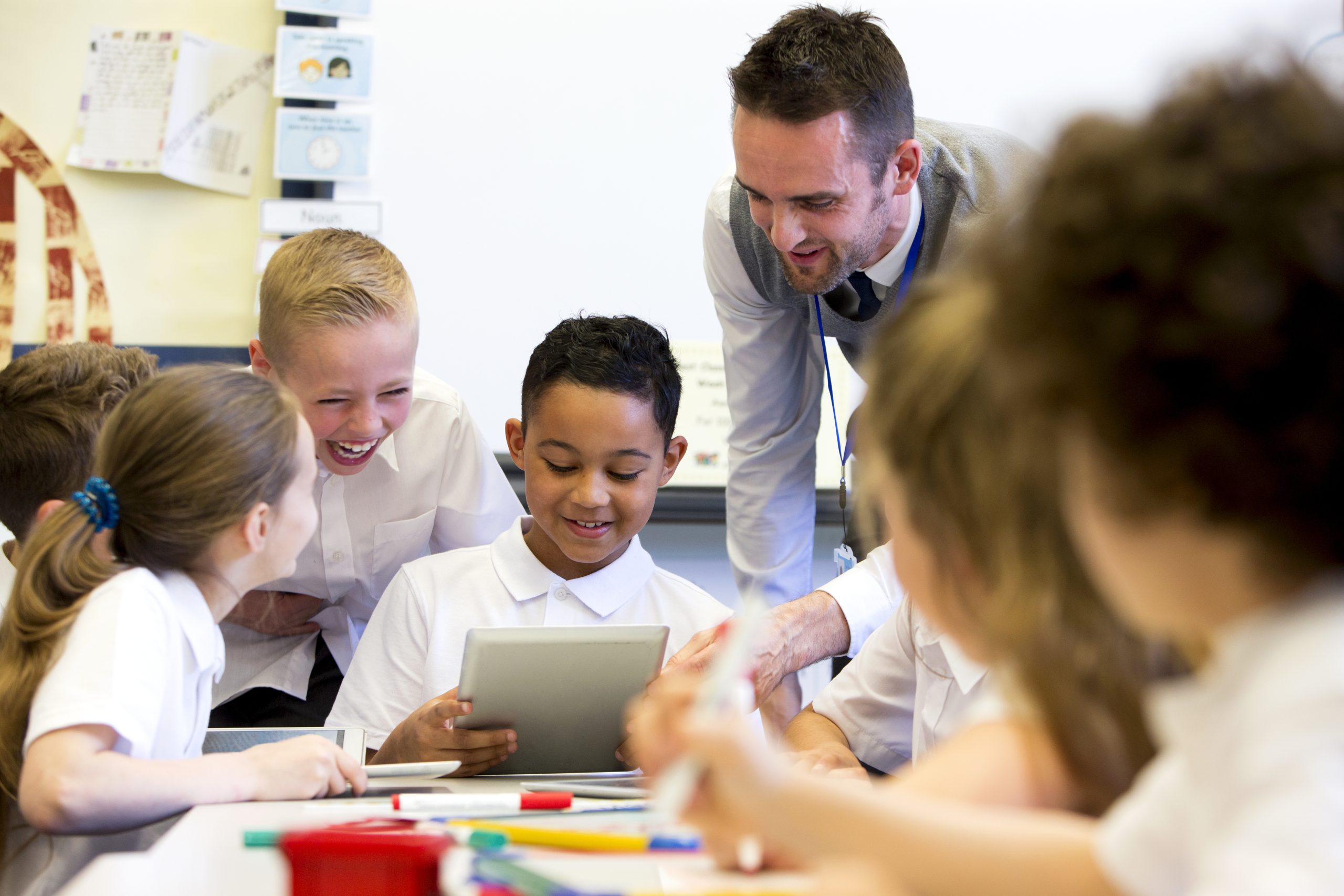 Photo of children wearing school uniform, sat at a table looking at an e-reader. There is an adult teacher behind them.