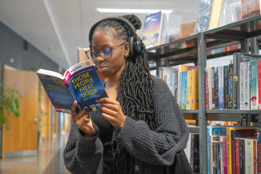 Photo of young woman stood in front of some book shelves, reading a Quick Reads book.